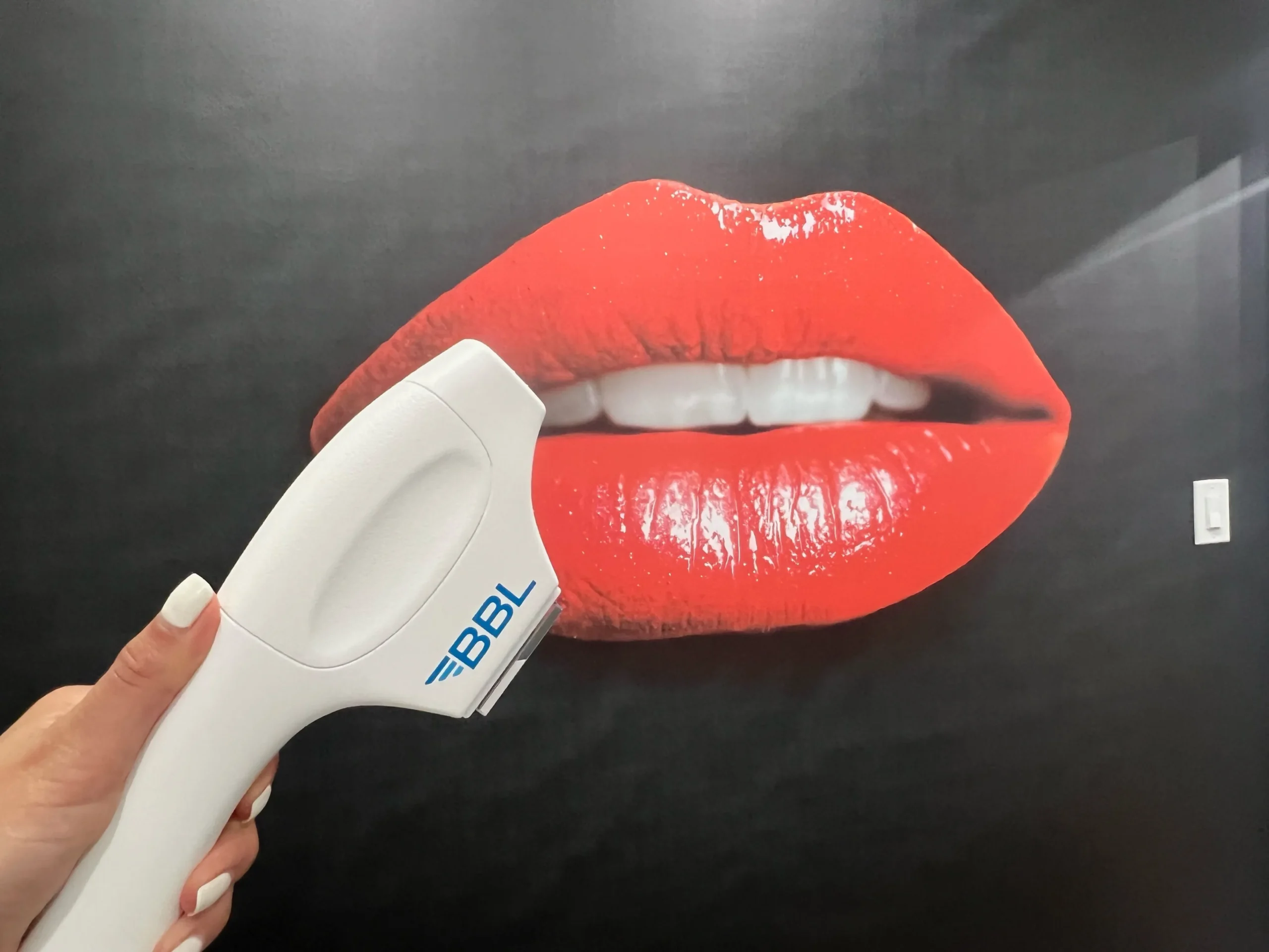 A hand holds a BBL Hero device in front of a red lip poster in Bellmore, NY