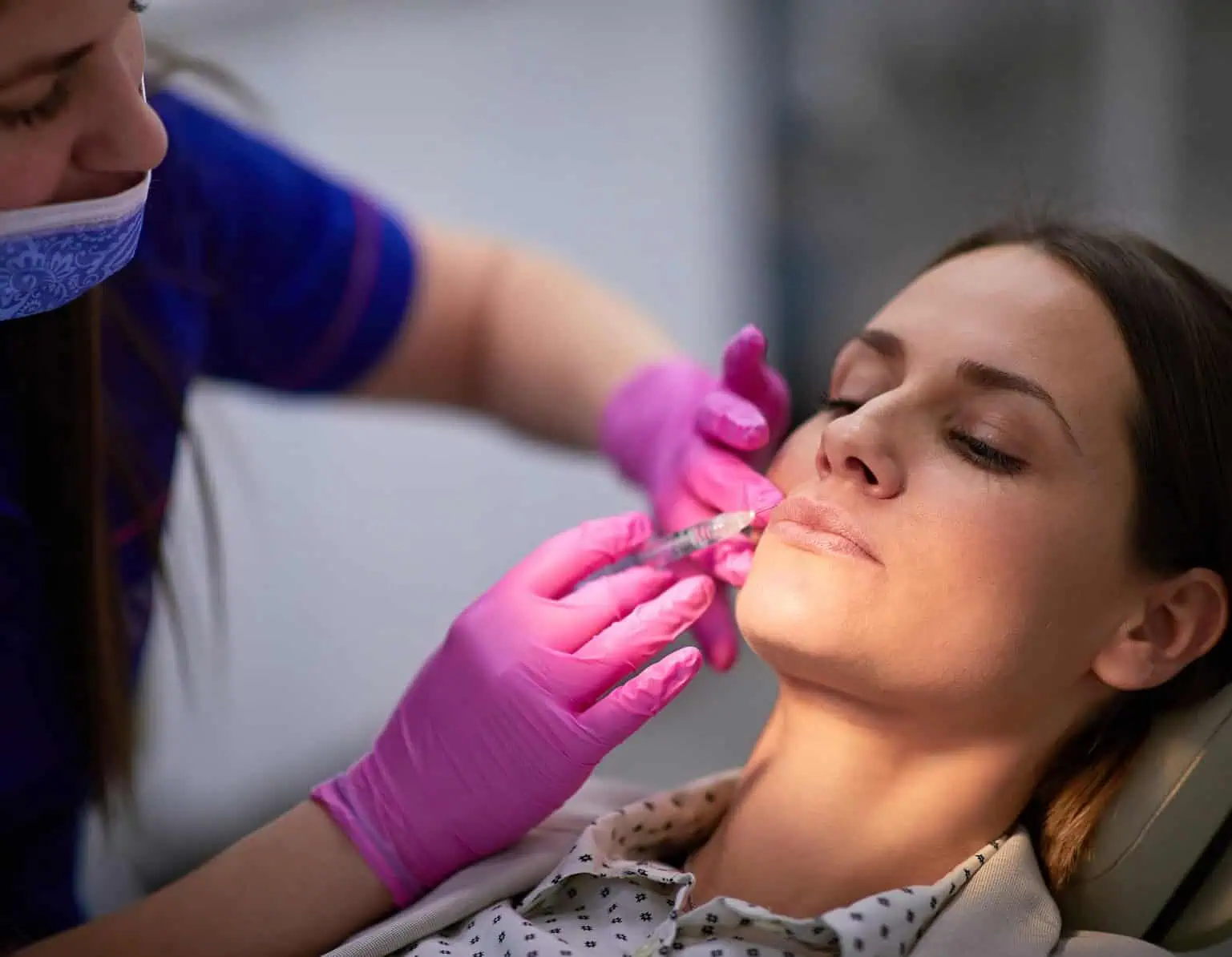 Medical professional in pink gloves administering cosmetic injectable treatment to woman's lips.